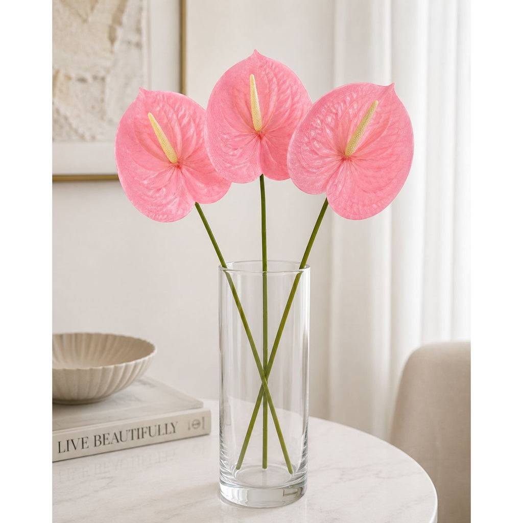 Three pink anthurium flowers in a clear glass vase on a white marble table, with a neutral indoor background.