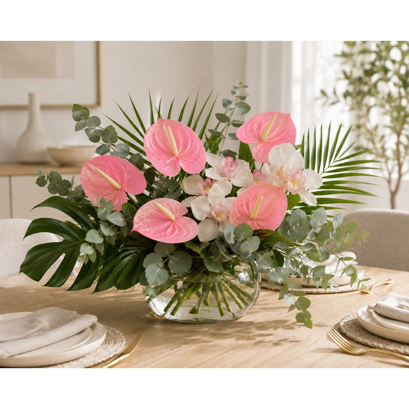 A vibrant pink and white floral arrangement featuring anthuriums, orchids, and green foliage in a clear glass vase, set on a light wood dining table with plates and chairs.