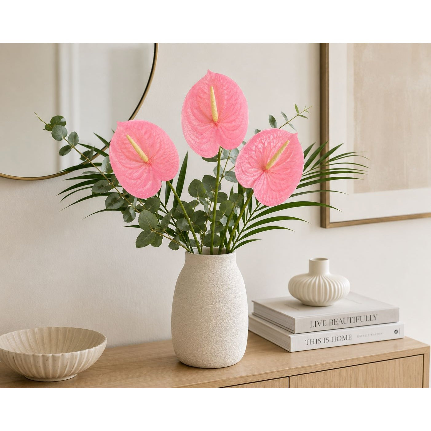 Three pink anthuriums in a textured cream ceramic vase, styled on a wooden console table with a bowl, books, and a small decorative vase in a bright, modern room.