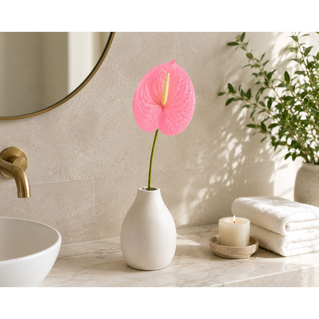 A pink anthurium flower in a matte white ceramic vase, styled on a marble bathroom counter next to a white sink, gold faucet, folded white towels, and a lit candle.