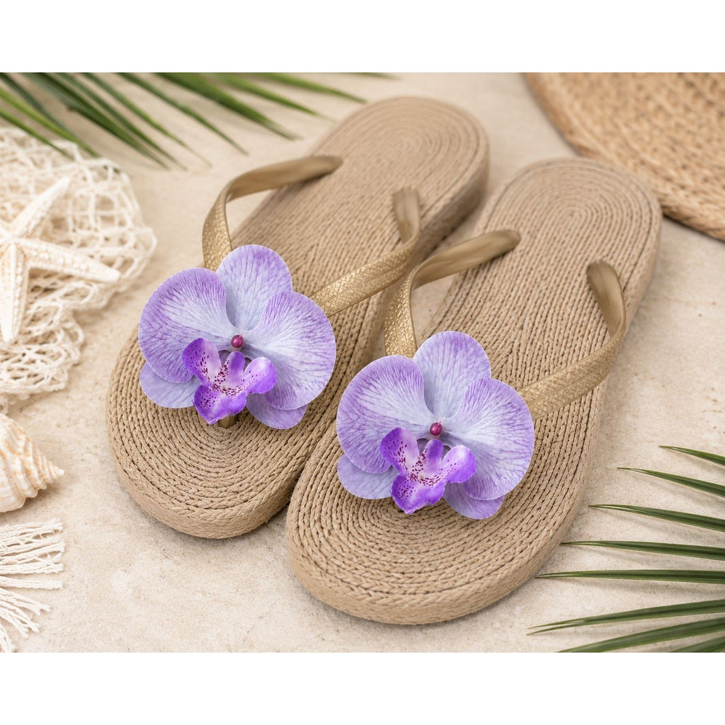 A pair of woven beige flip-flop sandals with large purple orchid flowers attached to the top, styled on a light sandy surface with tropical leaves and beach-themed props.