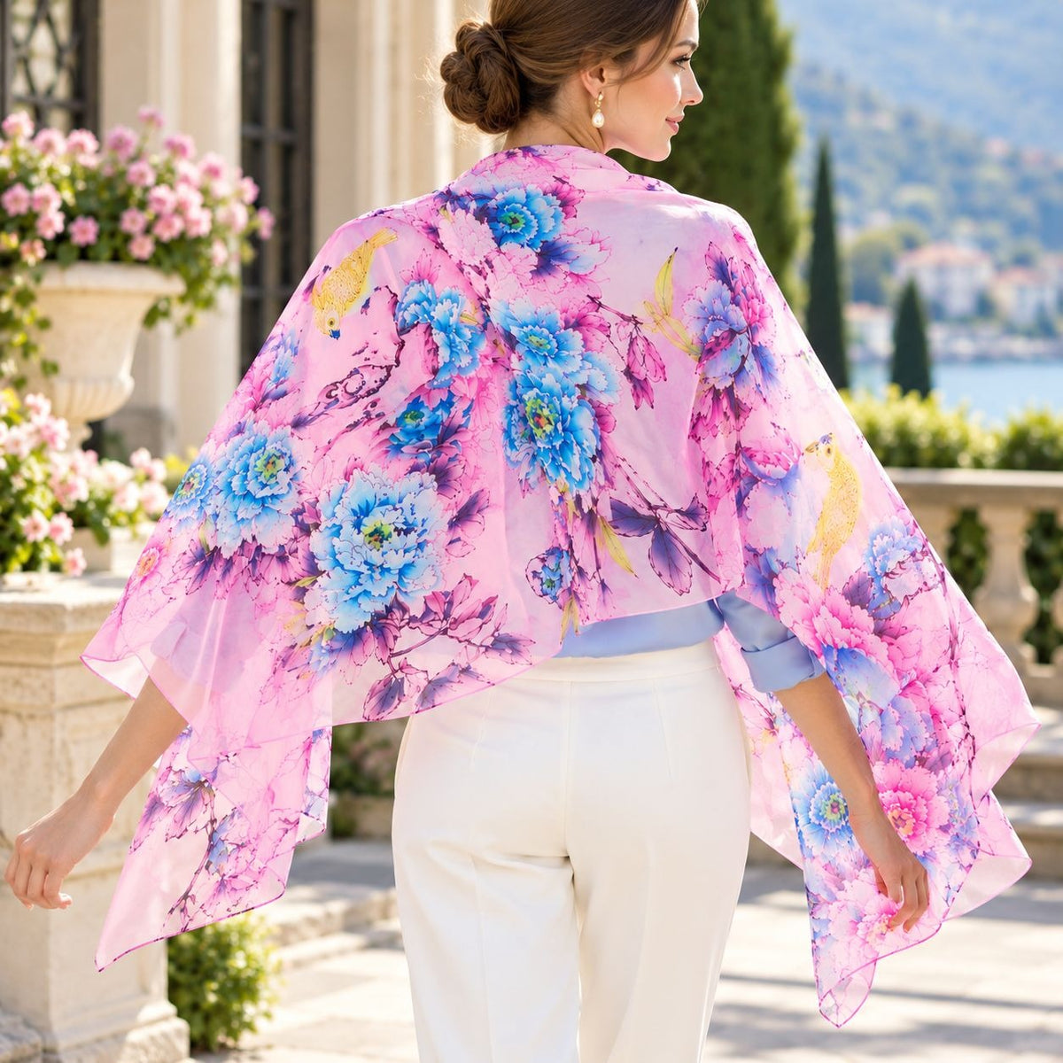 A woman wearing a sheer pink floral shawl with blue and yellow flowers, white pants, and a light blue top, photographed outdoors in a sunlit architectural setting.