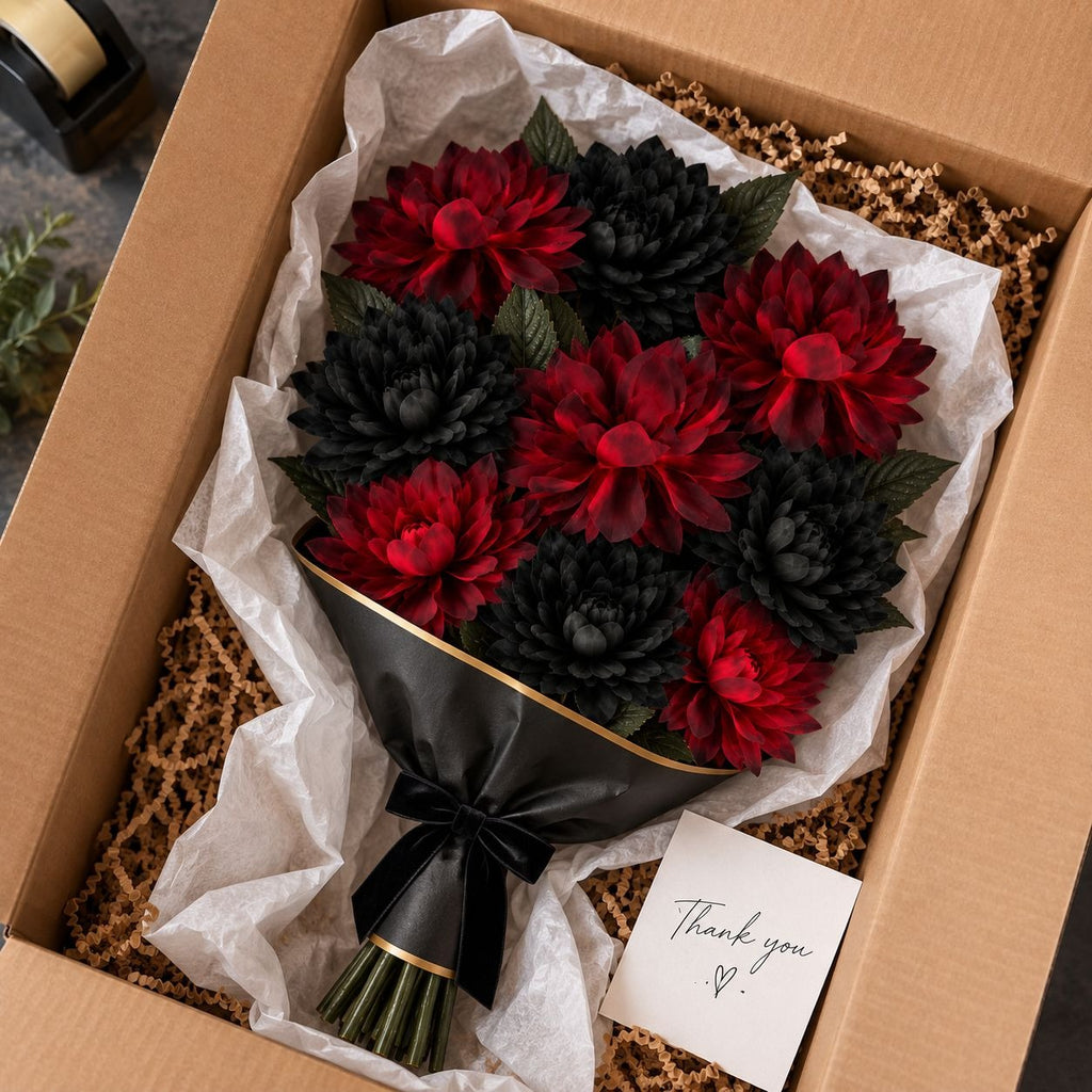 A bouquet of artificial red and black flowers wrapped in black paper with a gold band, presented inside a brown cardboard gift box with tissue paper and packing material. A small handwritten thank-you card is placed in the box.