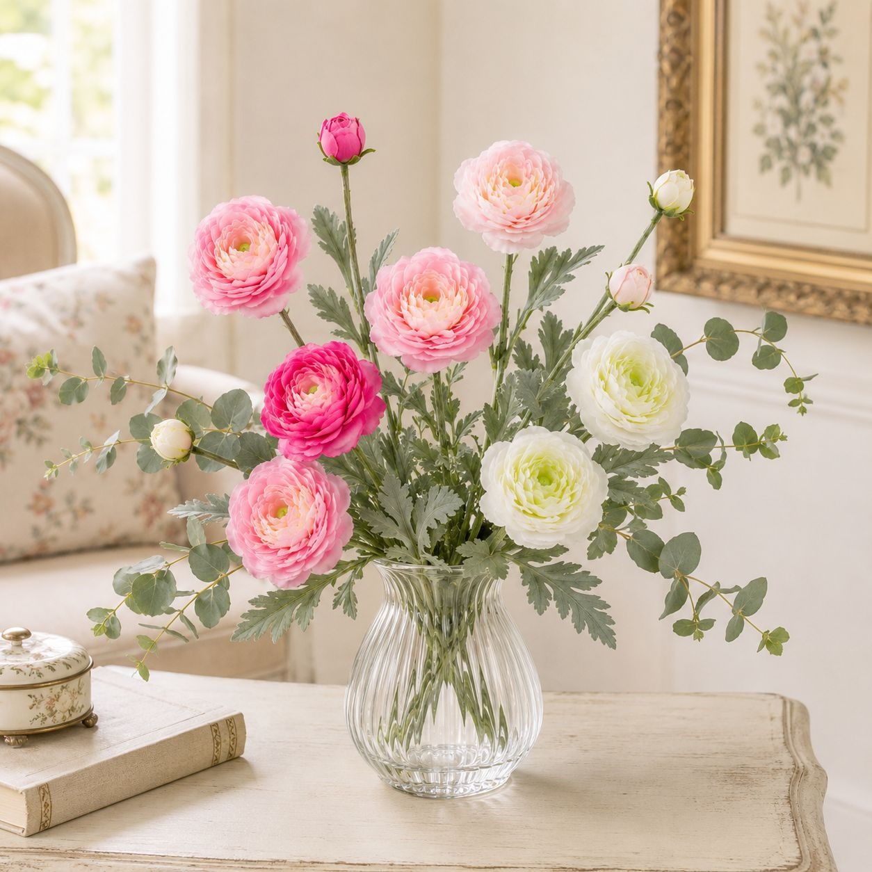 A floral arrangement of pink and white ranunculus flowers with green eucalyptus stems in a clear ribbed glass vase, placed on a light wood table in a softly lit interior setting.