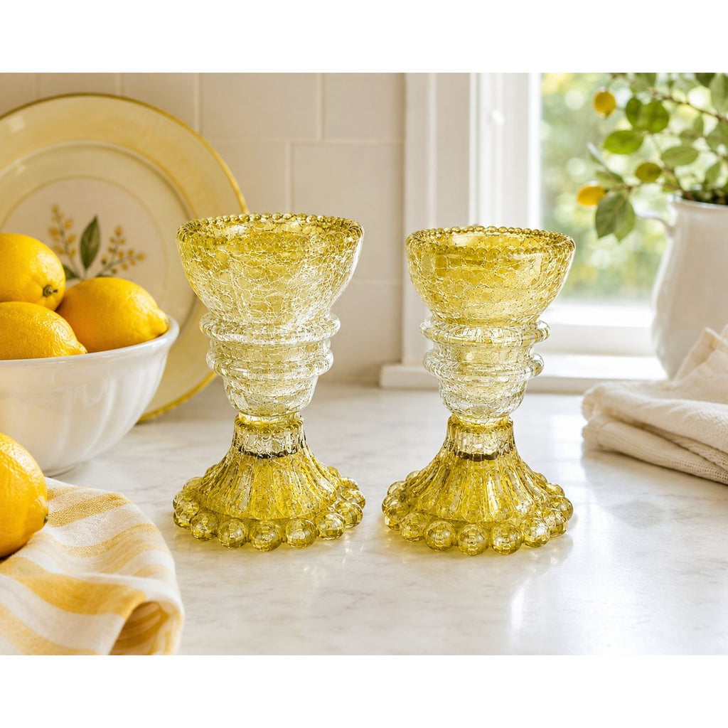 Two yellow crackle glass candleholders on a white marble countertop in a bright kitchen setting, with lemons, a plate, and a window in the background.