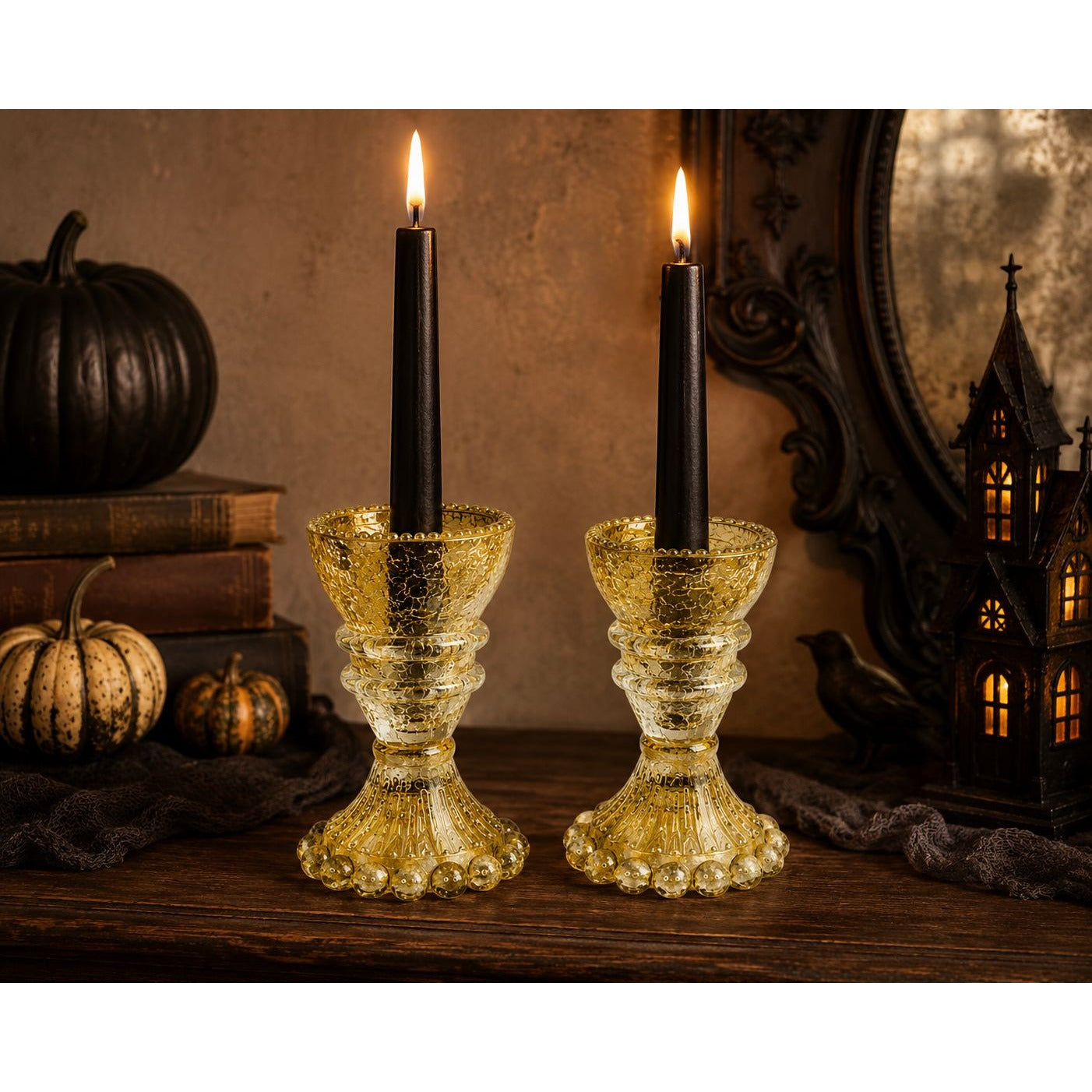 Two gold-toned textured glass candle holders with black taper candles, styled on a wooden surface with autumn decorations including pumpkins, books, and a lantern.