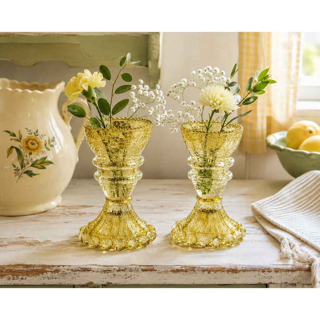Two small amber-yellow glass bud vases filled with white baby's breath and yellow flowers, displayed on a rustic white wooden table in a bright kitchen setting.