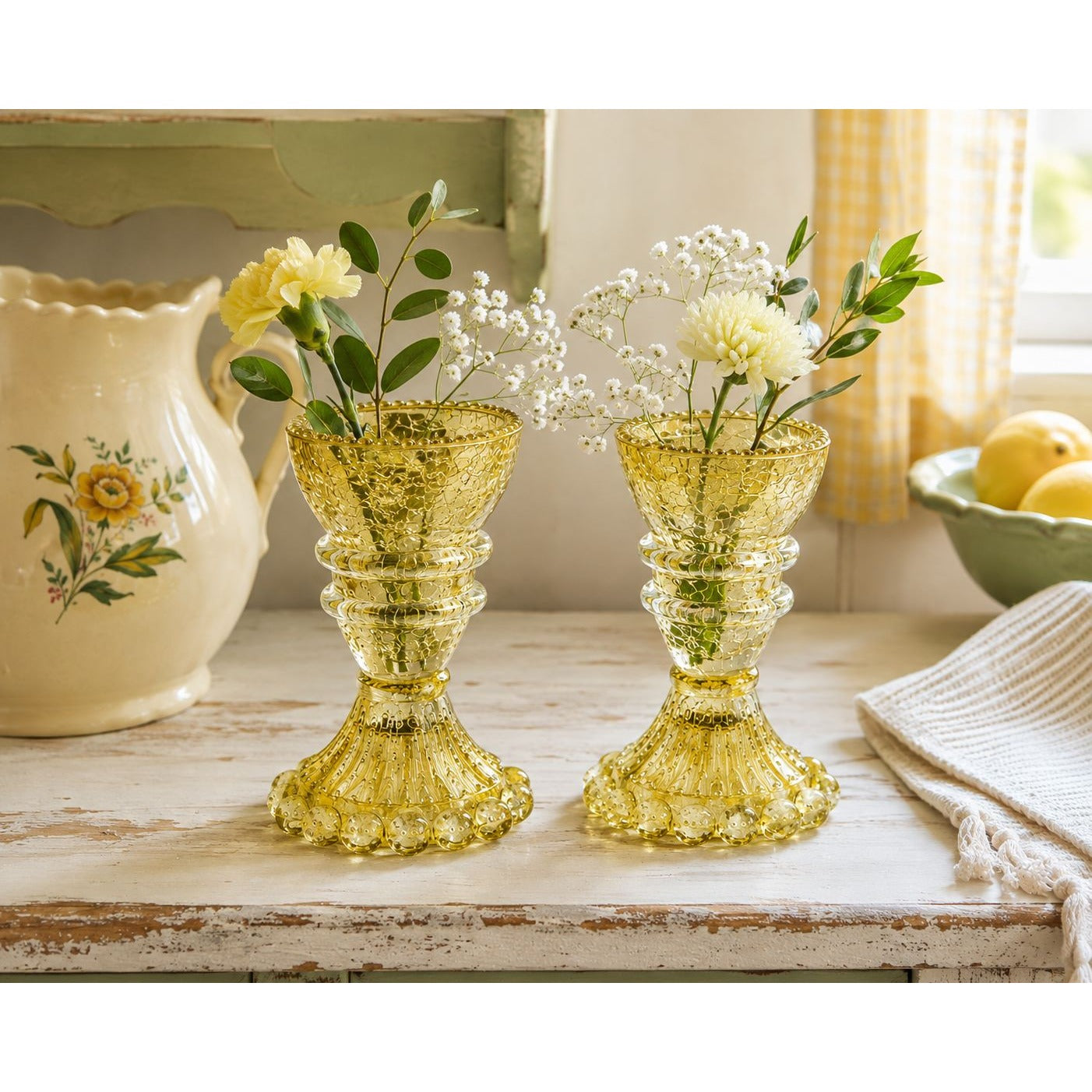 Two small amber-yellow glass bud vases filled with white baby's breath and yellow flowers, displayed on a rustic white wooden table in a bright kitchen setting.