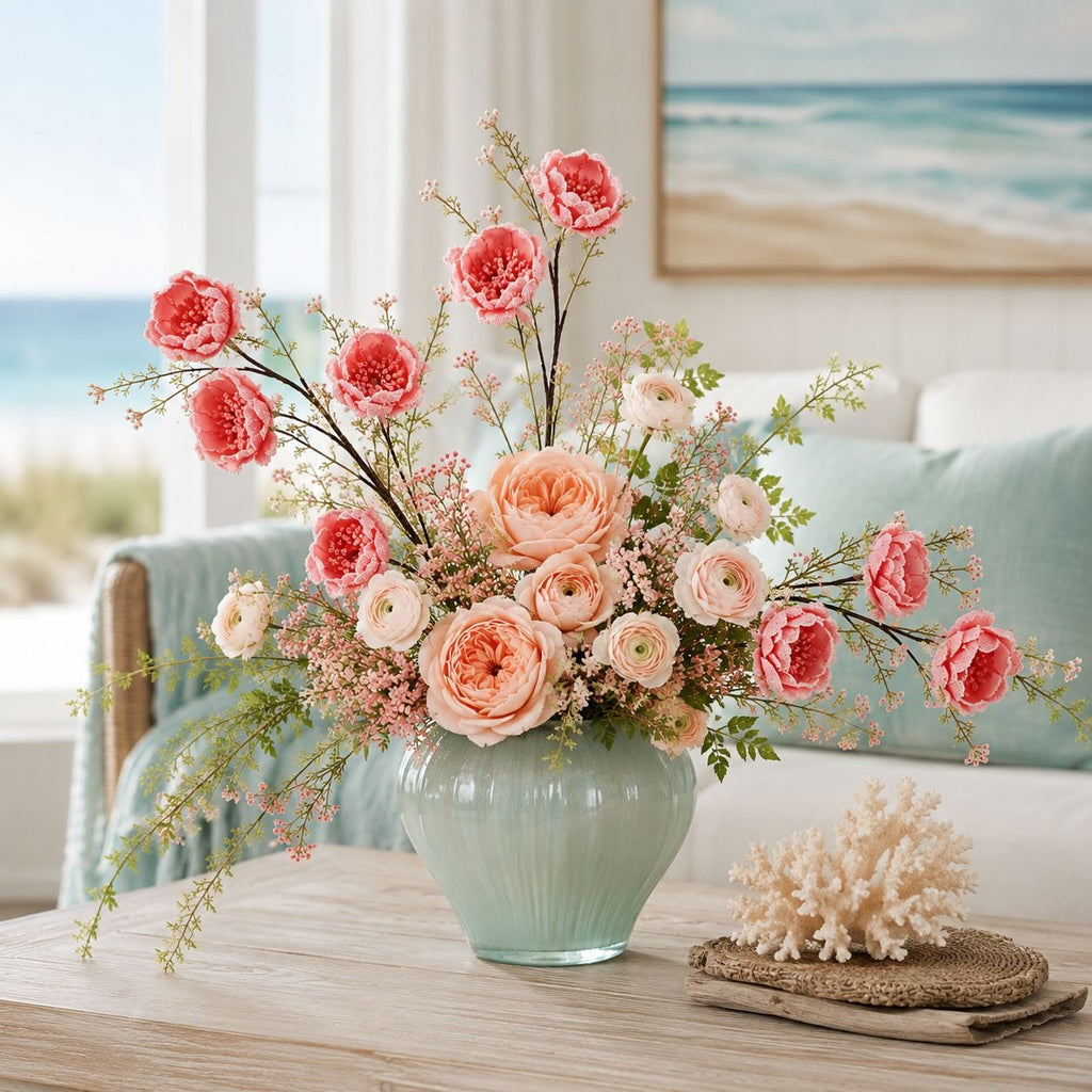 A floral arrangement in a pale aqua ribbed vase sits on a light wood table in a bright living room with a beach-view window and framed ocean artwork in the background. The bouquet includes coral-pink and peach roses, blush ranunculus, green foliage, and small filler flowers.