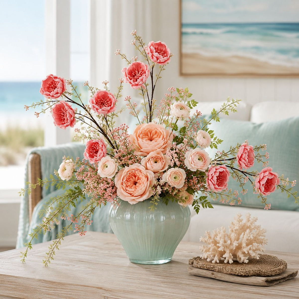A floral arrangement in a pale aqua ribbed vase sits on a light wood table in a bright living room with a beach-view window and framed ocean artwork in the background. The bouquet includes coral-pink and peach roses, blush ranunculus, green foliage, and small filler flowers.