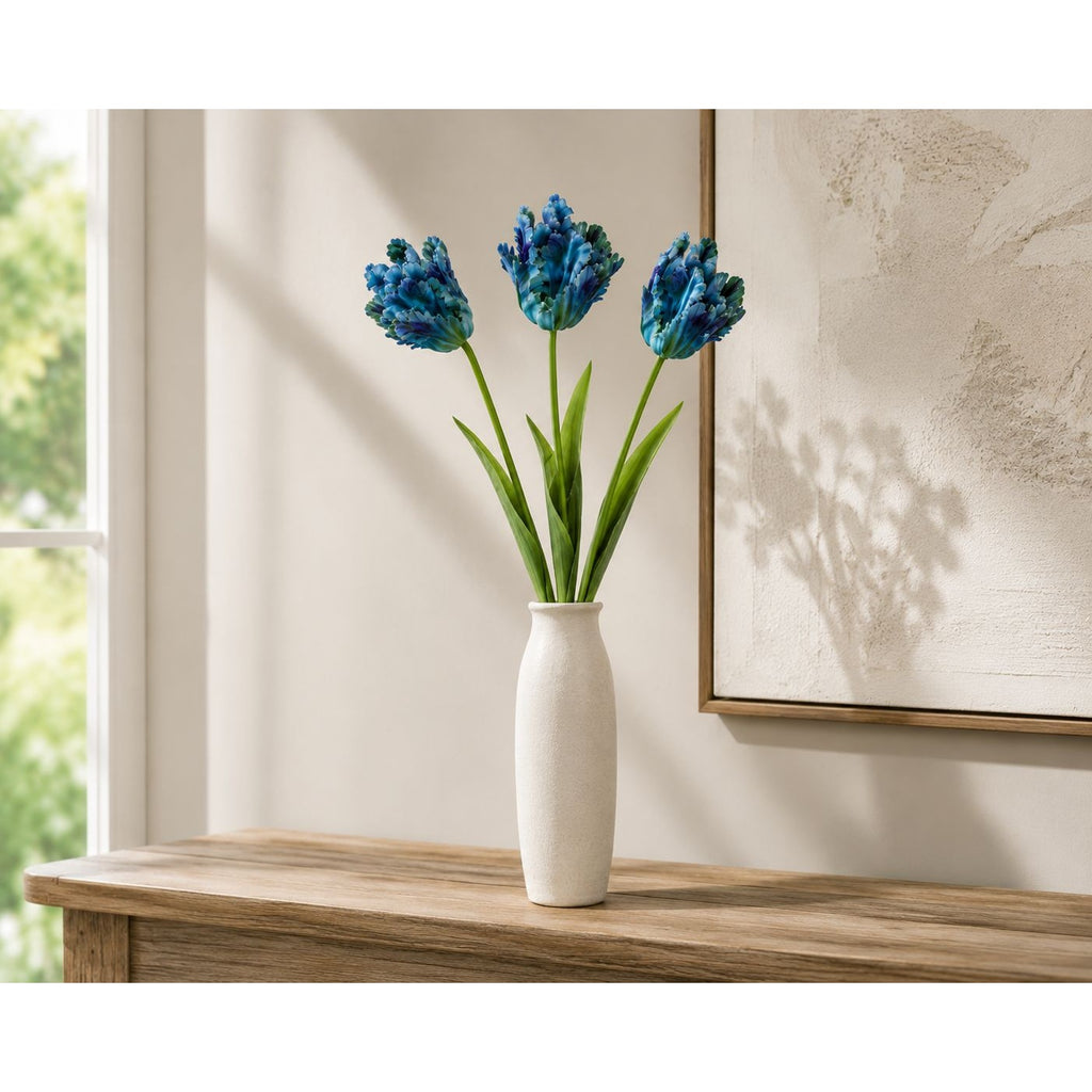Three blue tulip flowers in a white ceramic vase, displayed on a wooden console table against a neutral wall with a framed print.