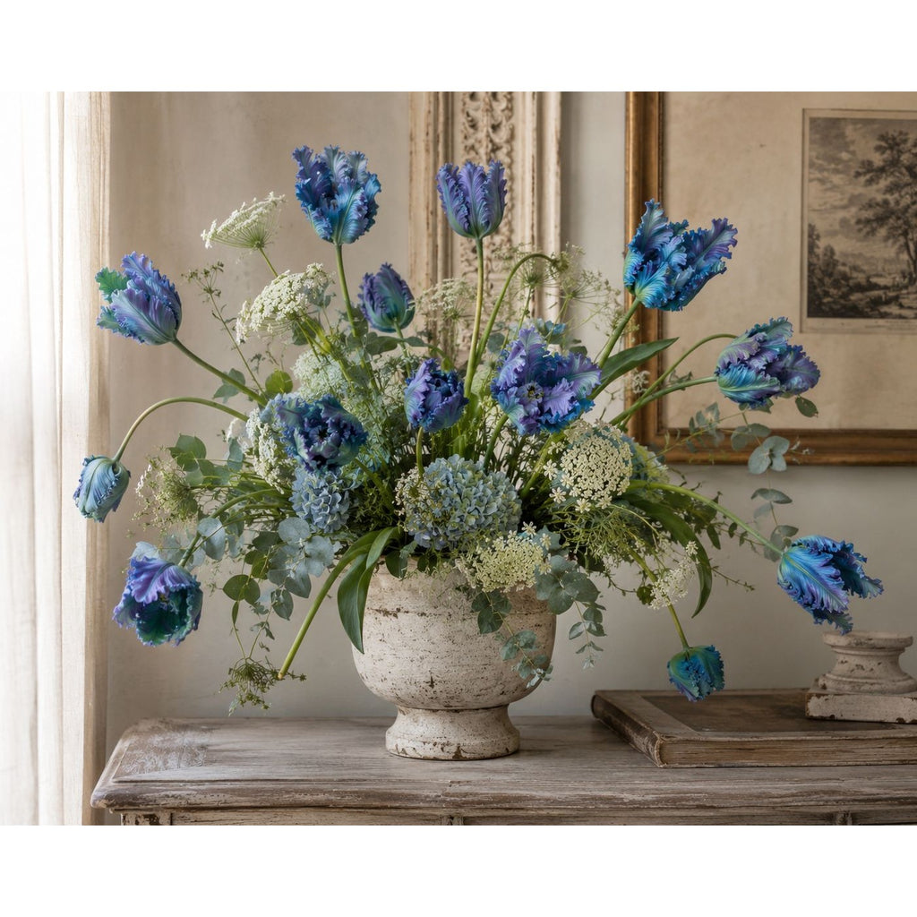A large floral arrangement of blue tulips,  and green foliage in a distressed white ceramic urn, displayed on a rustic wooden console table in an elegant room.