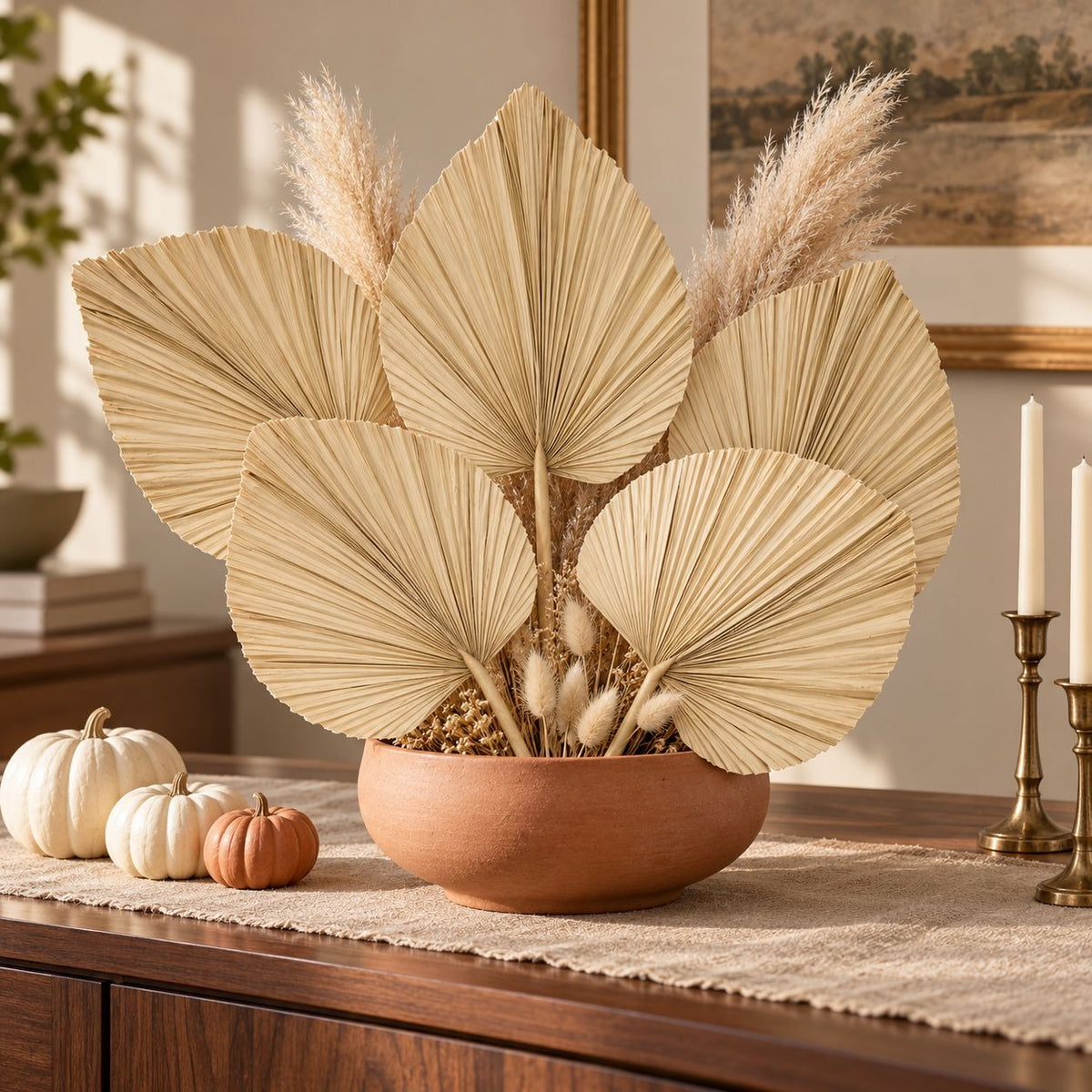A decorative arrangement of large beige dried palm leaves and pampas grass in a round terracotta bowl, displayed on a wooden table with small pumpkins and candles in a warm indoor setting.