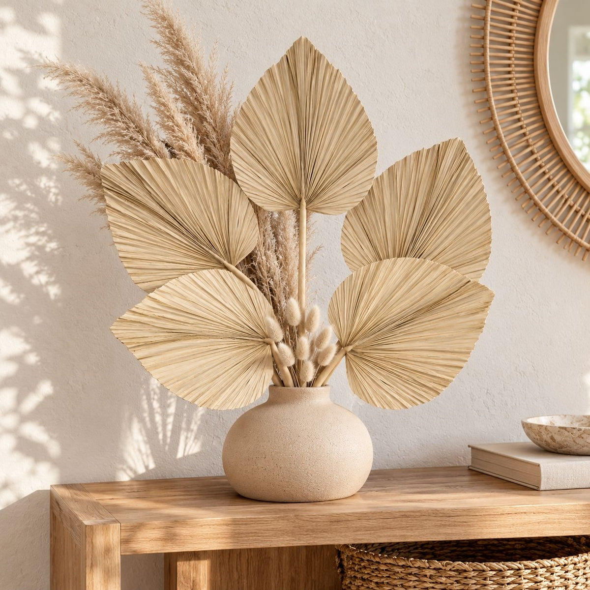 A decorative arrangement of dried palm-leaf and pampas-style stems in a round beige ceramic vase, displayed on a wooden console table in a warm neutral interior.