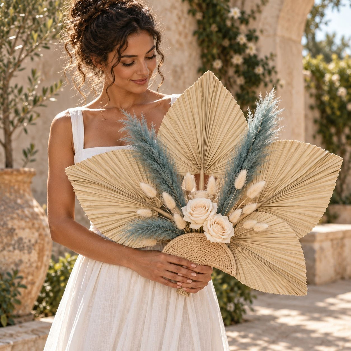 A woman in a white sleeveless dress holds a large decorative dried flower arrangement featuring tan fan palm leaves, blue pampas grass, cream roses, and a woven basket base, photographed outdoors in a sunlit stone courtyard.