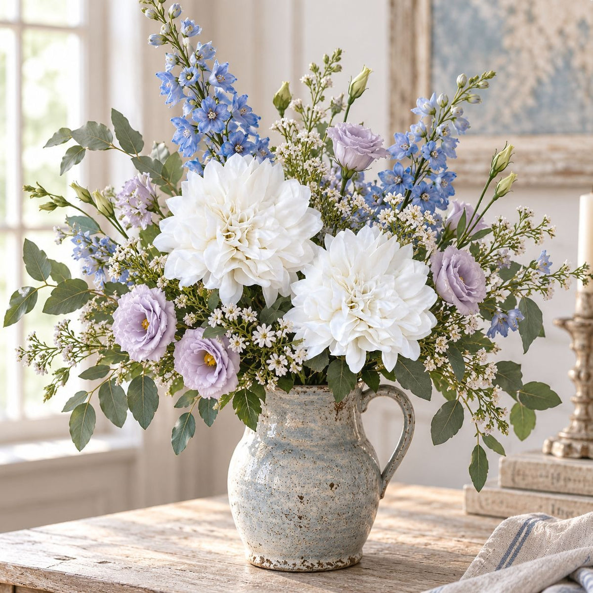 A rustic ceramic pitcher vase filled with a floral arrangement of white dahlias, light purple roses, blue delphiniums, and green foliage, set on a wooden table in a bright room.