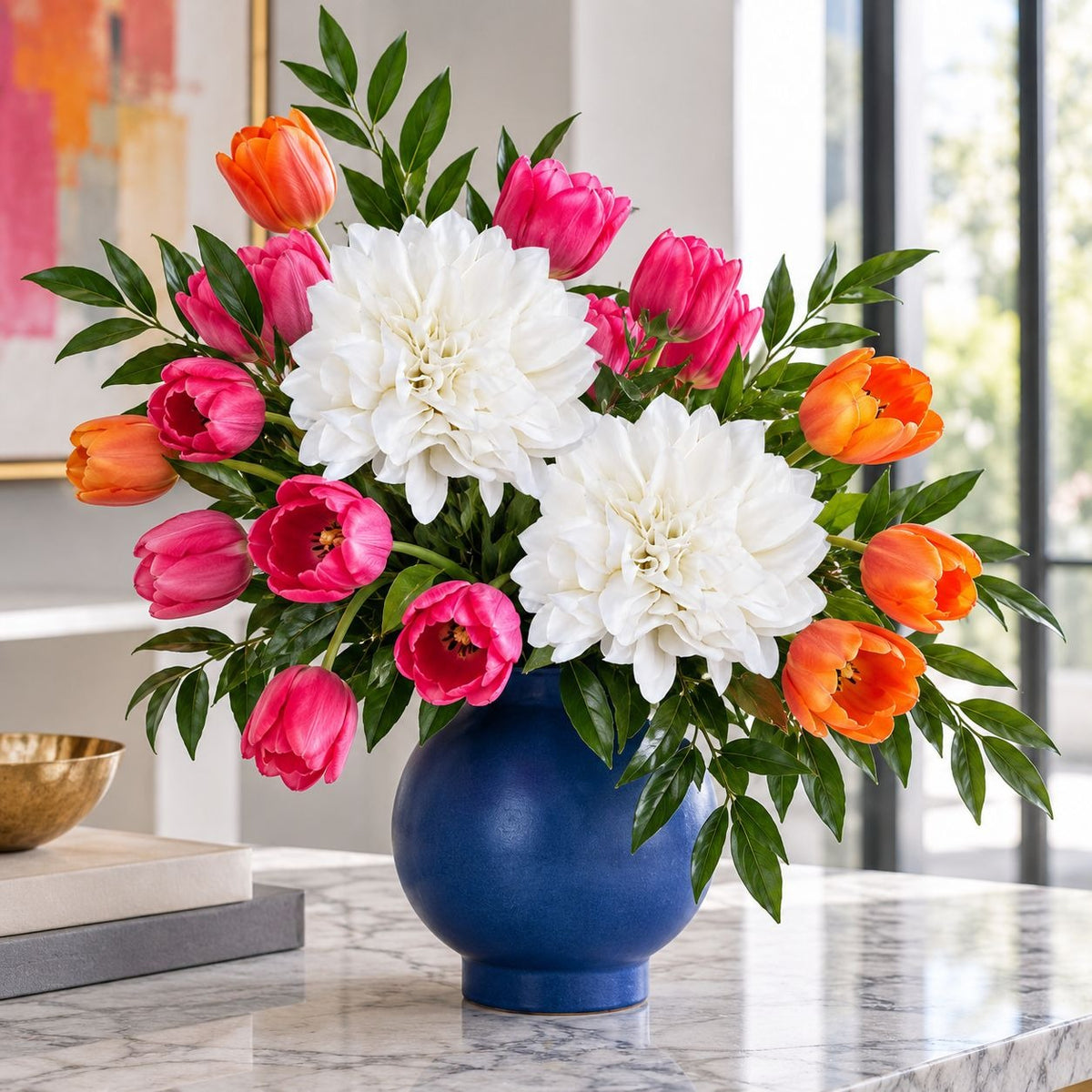 A vibrant artificial flower arrangement featuring large white dahlias, bright pink tulips, and orange tulips with green leaves, presented in a glossy dark blue ceramic vase on a marble countertop.