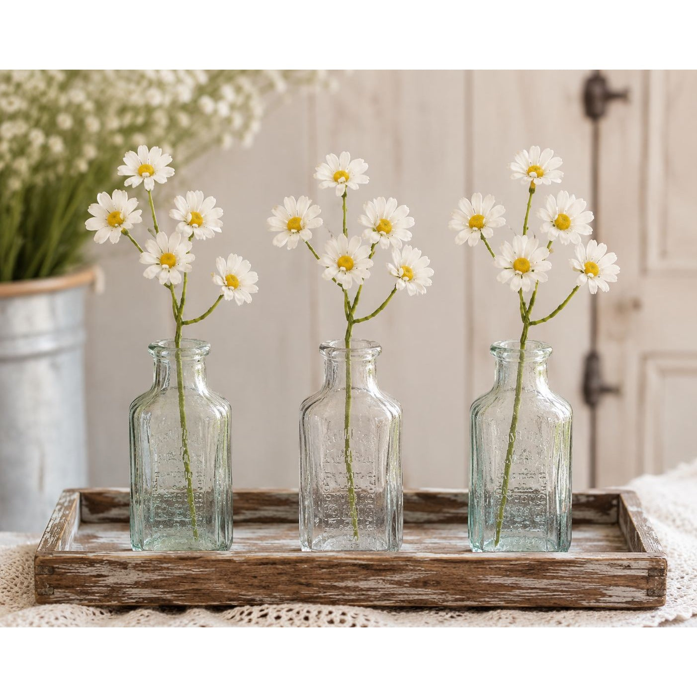 Three clear glass bottles with daisies on a wooden tray in a home setting.