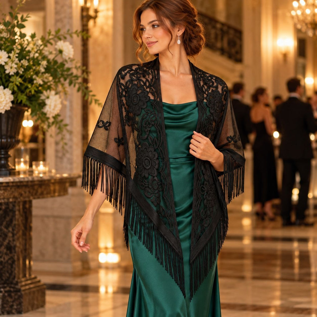 A woman in a formal indoor setting wears a black lace and fringe shawl over a dark green satin gown. The background shows a softly blurred elegant hall with warm lighting and other guests.