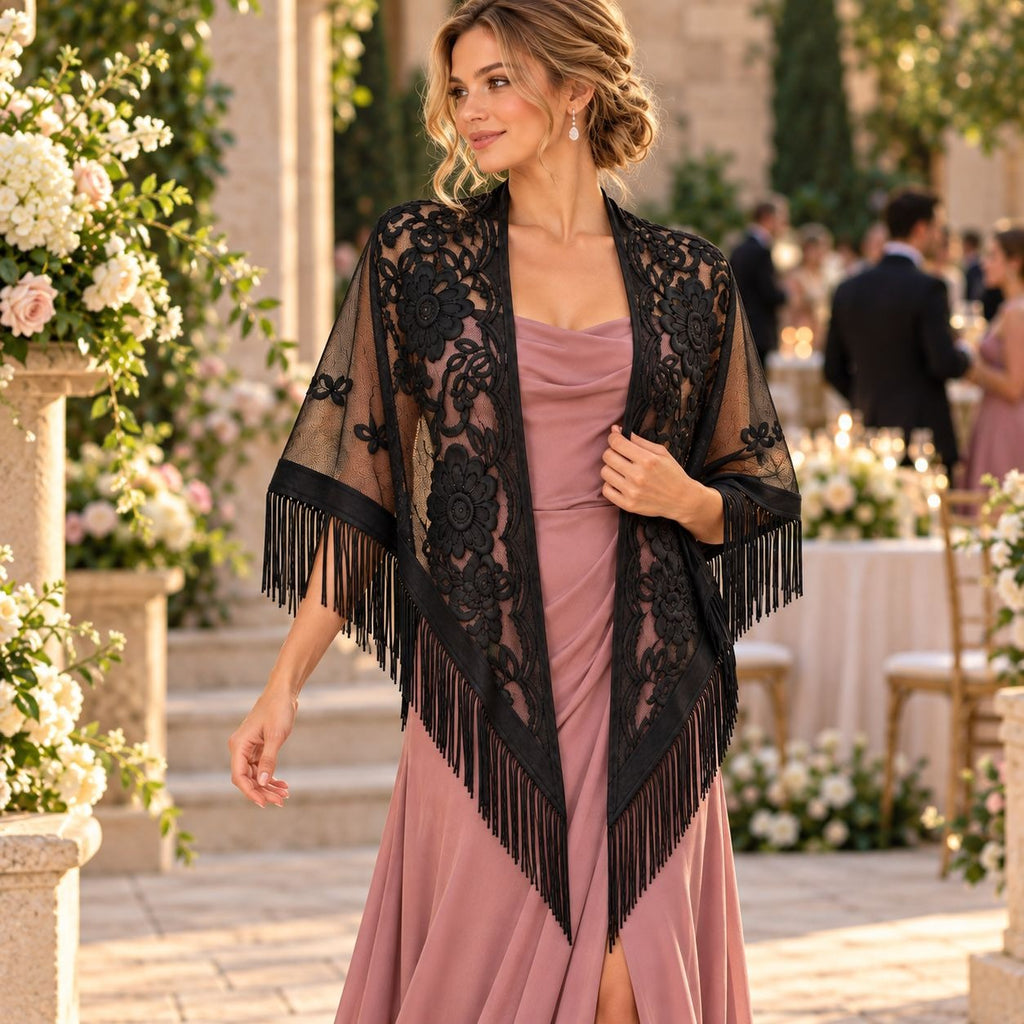 A woman stands in an outdoor garden or event setting wearing a black lace and fringe shawl over a dusty pink strapless dress. The background shows blurred tables, flowers, and guests.