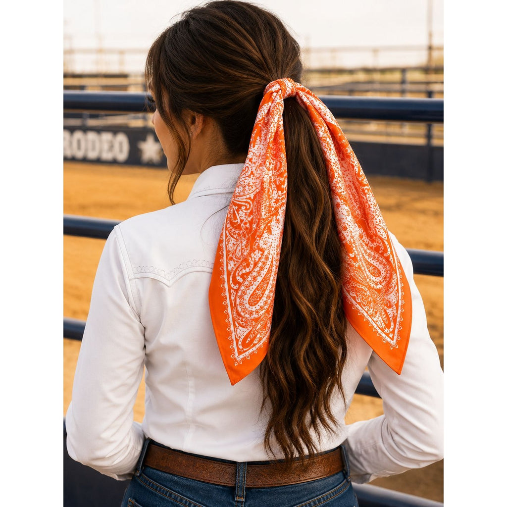 A woman seen from behind wearing a white western-style shirt and blue jeans, with a bright orange paisley hair scarf tied around her brown ponytail in an outdoor rodeo arena.