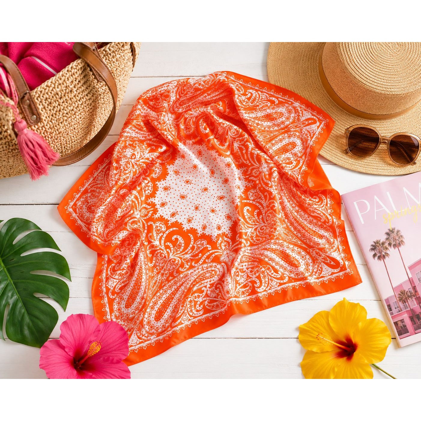 An orange and white paisley bandana scarf is styled in a flat lay on a white wooden surface, surrounded by tropical flowers, a straw hat, sunglasses, a woven tote bag, and a pink magazine.