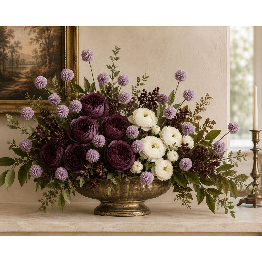 A large bronze pedestal bowl filled with an elegant floral arrangement featuring deep burgundy roses, white ranunculus, lavender globe flowers, and green foliage, set on a stone ledge in a classic interior setting.