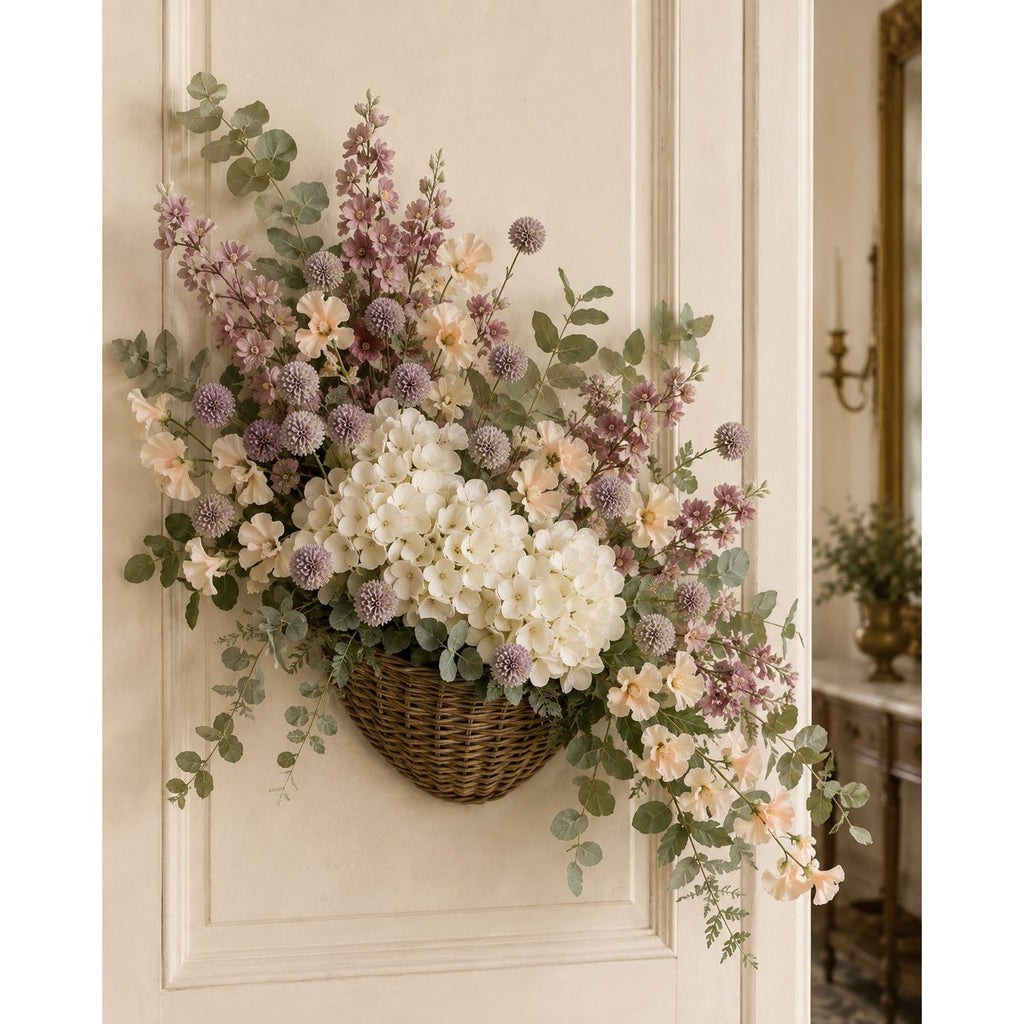 A hanging wicker basket filled with a floral arrangement of white hydrangeas, dusty purple pom-pom flowers, and green eucalyptus leaves, displayed on a cream-colored paneled door.