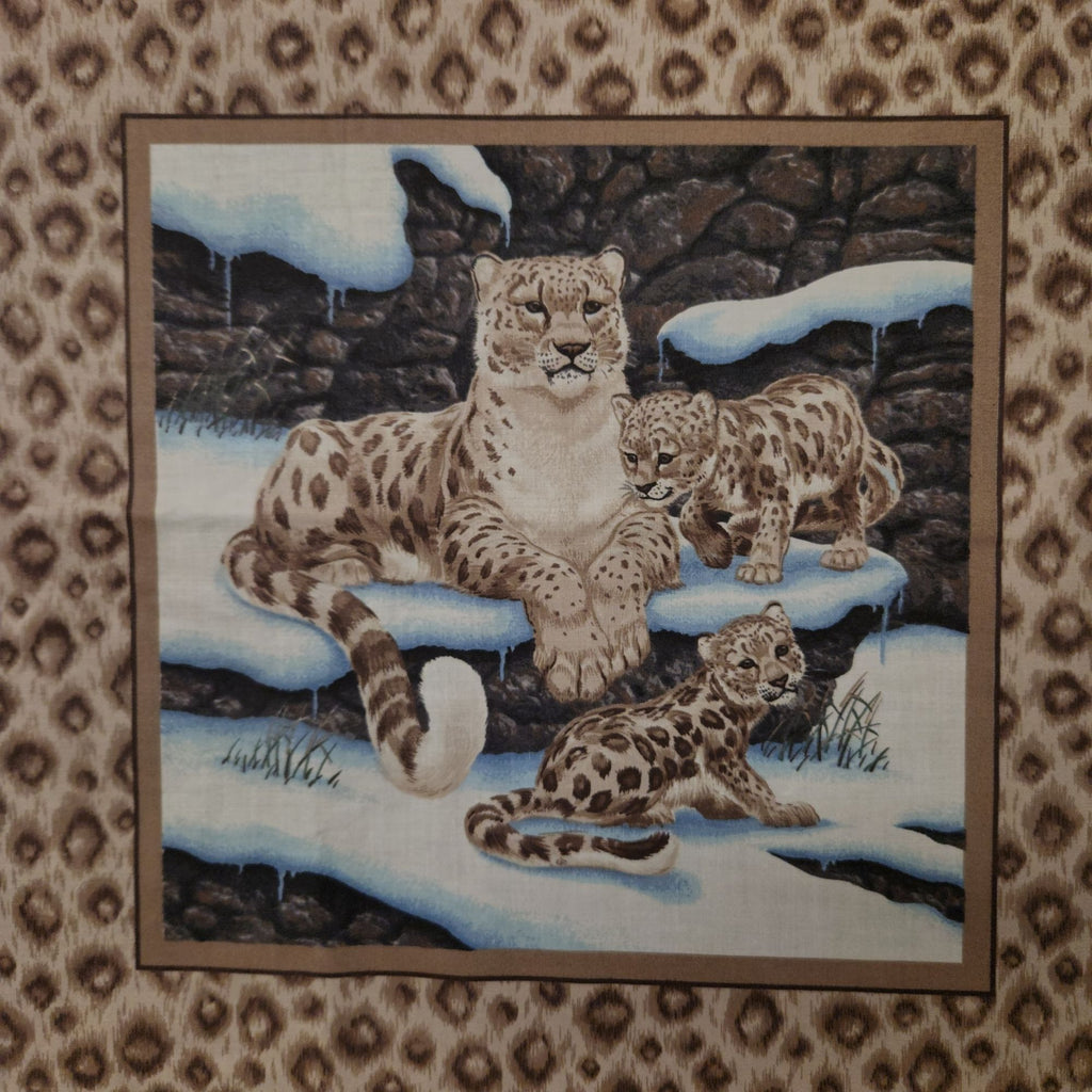 A square textile product, likely a tapestry or wall hanging, featuring a detailed illustration of three snow leopard cubs in a snowy, rocky outdoor setting. The tapestry is framed by a brown border and is displayed flat against a matching leopard-print fabric background.