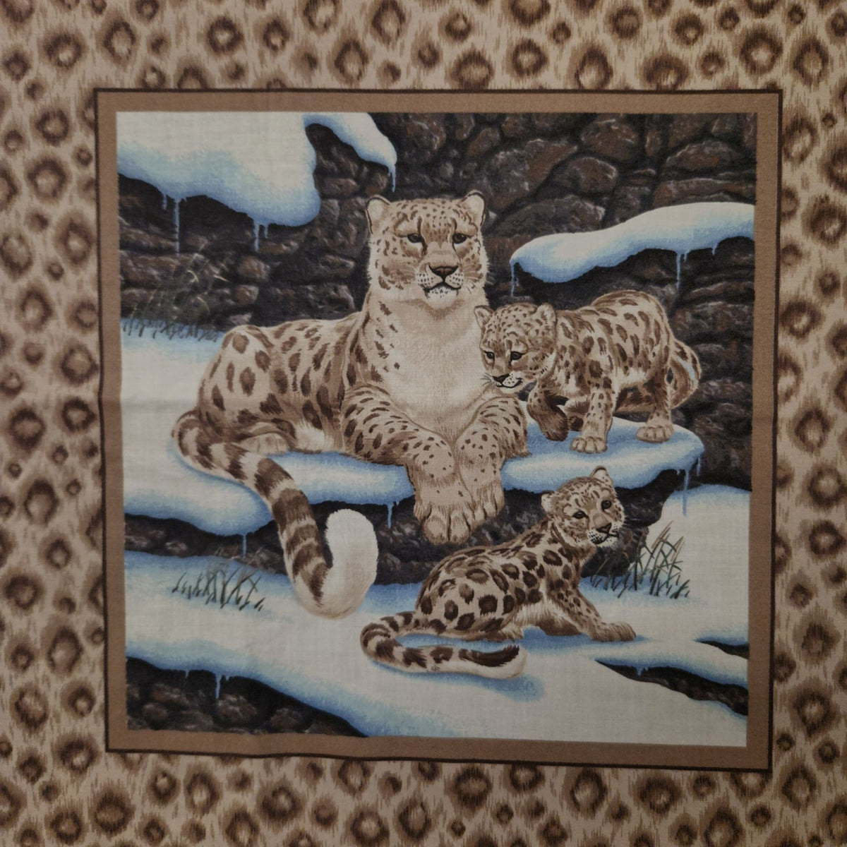 A square textile product, likely a tapestry or wall hanging, featuring a detailed illustration of three snow leopard cubs in a snowy, rocky outdoor setting. The tapestry is framed by a brown border and is displayed flat against a matching leopard-print fabric background.