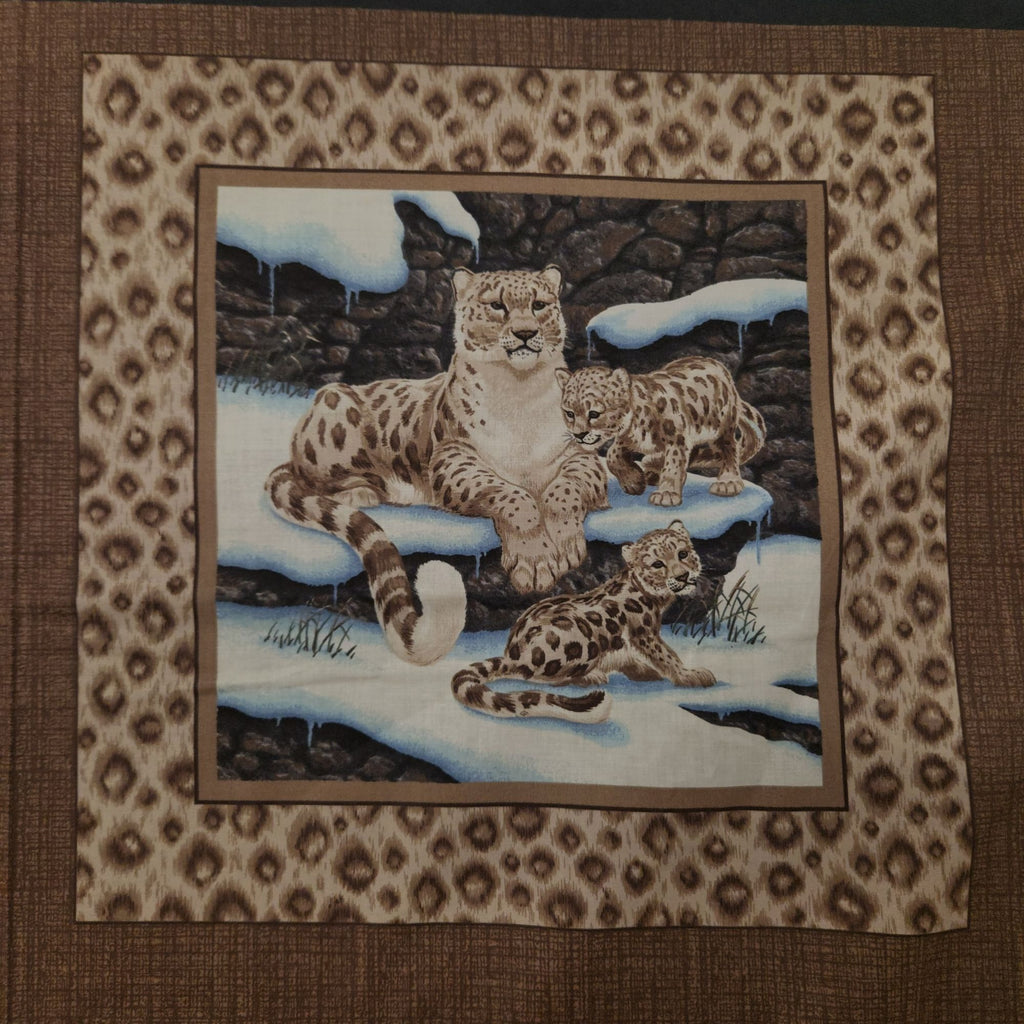 A square textile or wall hanging featuring a central painted scene of three snow leopard cats in a snowy rocky setting, surrounded by a wide brown patterned border on a tan background.