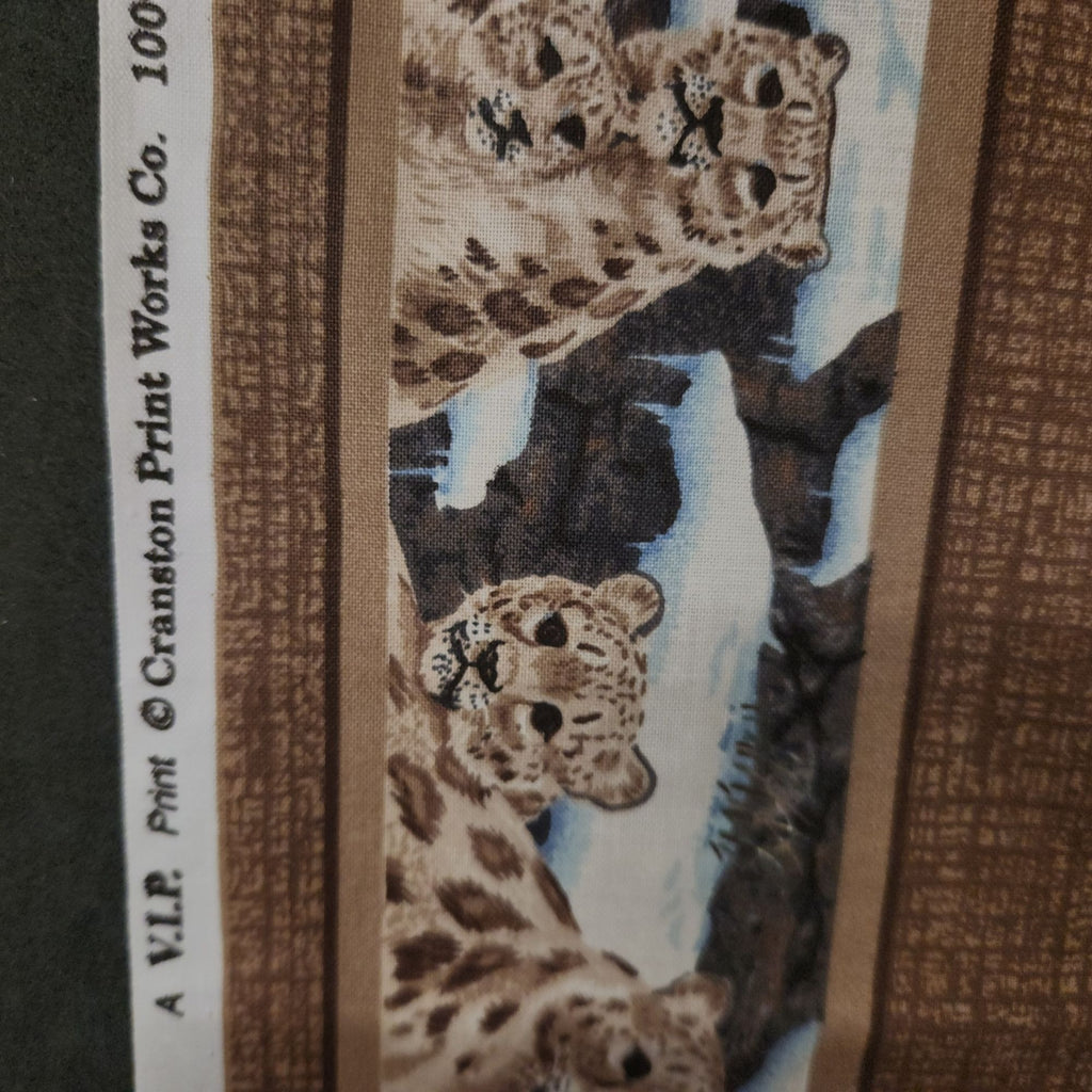 A close-up of a printed textile or wallpaper sample featuring three spotted big cats in a snowy mountain scene, with brown patterned side borders and vertical text reading 'A V.I.