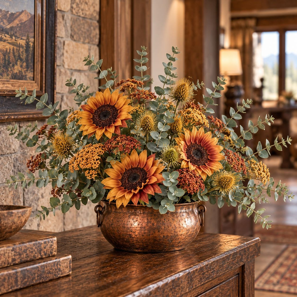 A copper flower arrangement bowl filled with sunflowers, eucalyptus, and autumnal filler flowers, displayed on a wooden console table in a warm, rustic interior setting.
