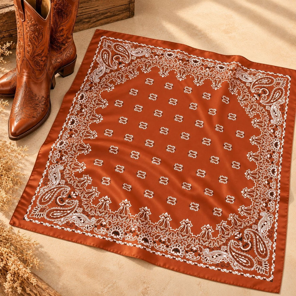 A rust-orange bandana with a white paisley border and repeating monogram pattern, styled flat on a beige surface next to brown cowboy boots.