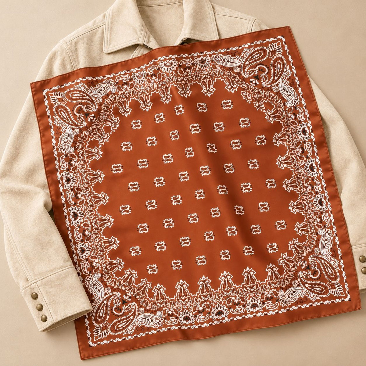 A rust-orange bandana with a white paisley border and small repeating motifs, laid flat over a cream-colored collared shirt on a beige background.