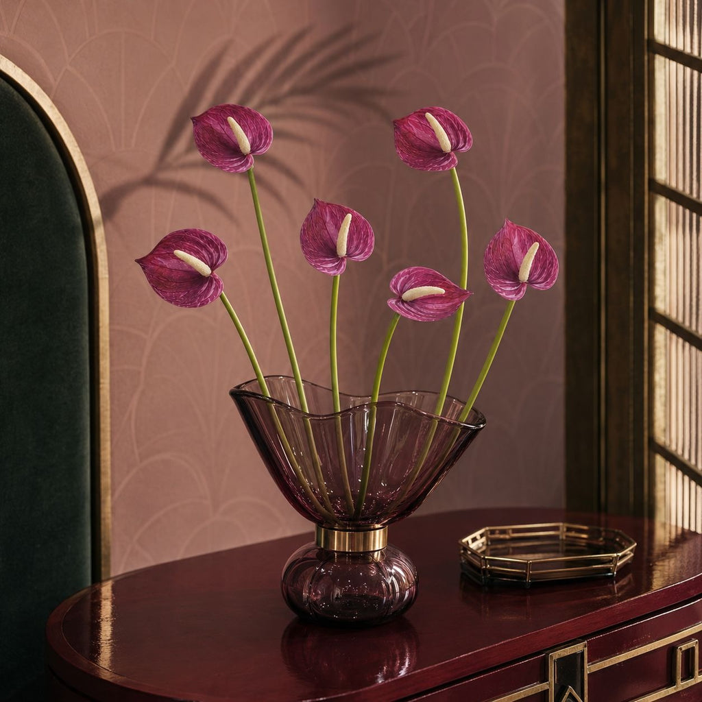 A purple glass vase with a flared bowl and rounded base, holding several pinkish-purple anthuriums with long green stems, placed on a dark wooden table in an interior setting with patterned wallpaper and a window.