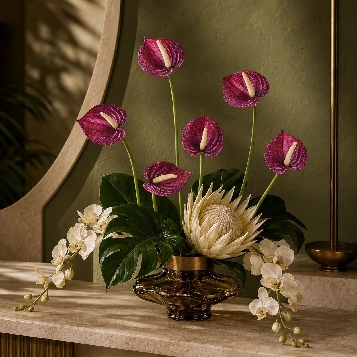 A floral arrangement in a dark amber glass vase on a stone console table, featuring magenta anthuriums, a cream protea, white orchids, and green foliage against an olive-green wall.