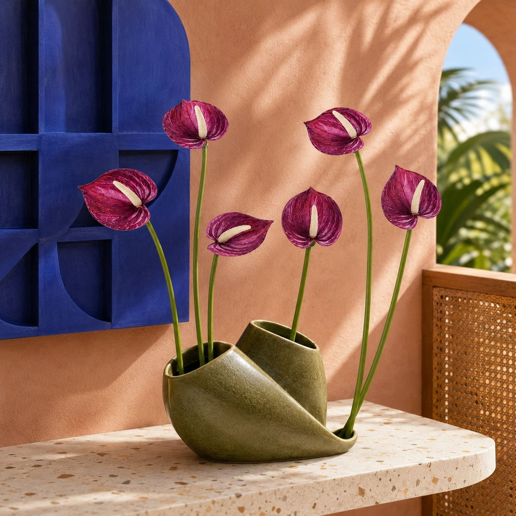 A sculptural olive green ceramic vase holding five purple anthurium flowers, styled on a speckled stone ledge against a warm terracotta wall with a blue geometric screen and outdoor greenery in the background.