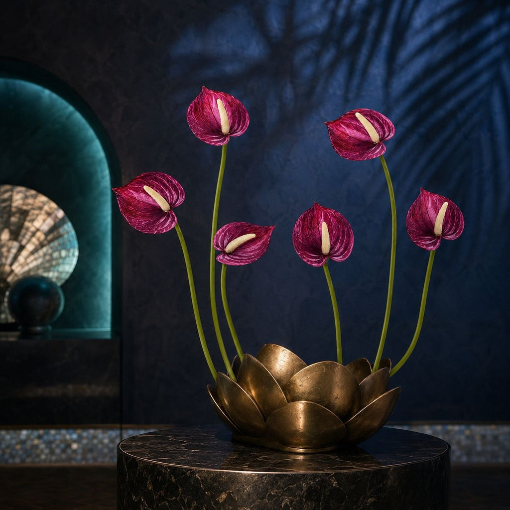 A decorative floral arrangement featuring six magenta-pink anthuriums on long green stems, displayed in a gold lotus-shaped vase on a dark marble pedestal against a dark blue wall with shadow patterns.