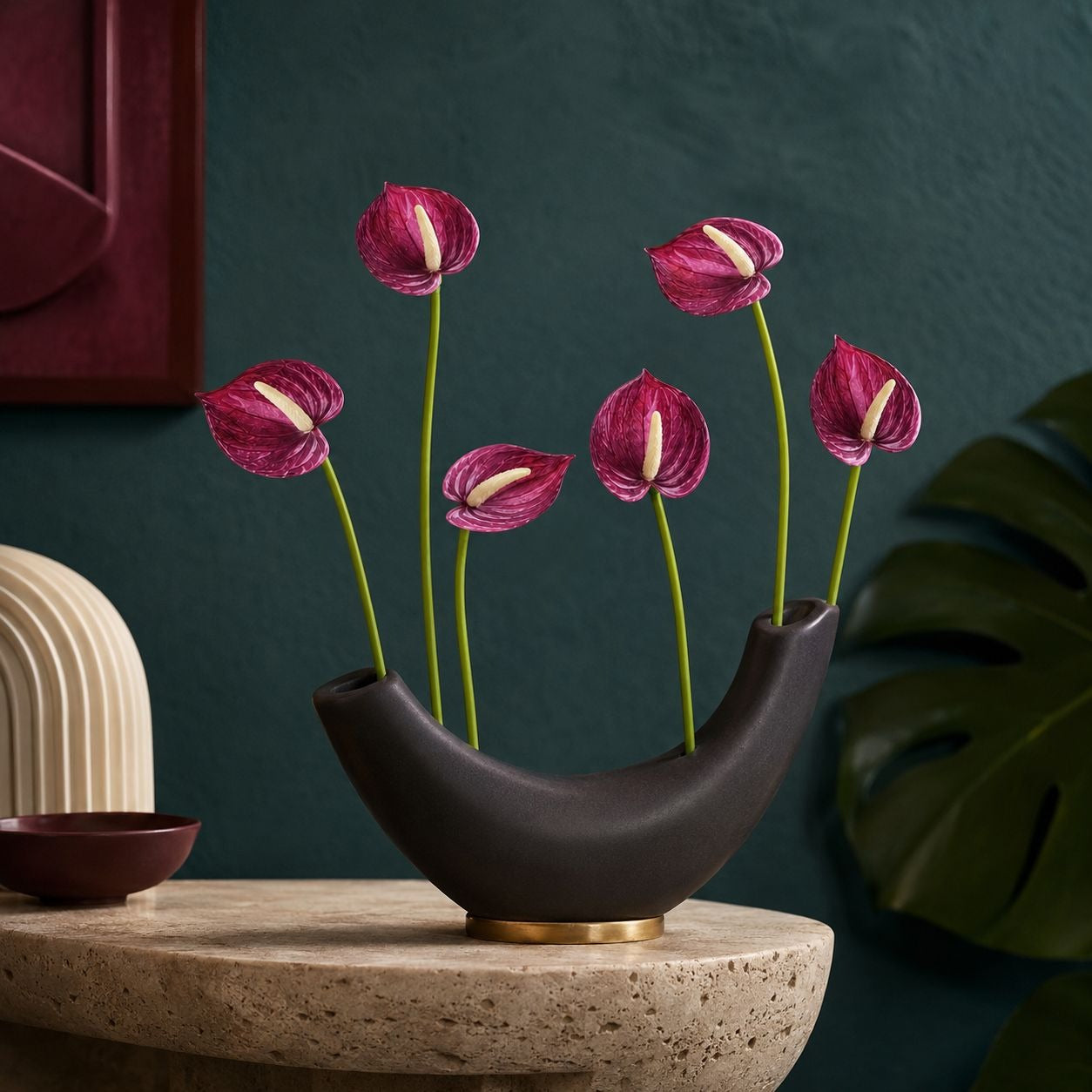 A dark brown crescent-shaped ceramic vase with a gold base accent, holding six pinkish-purple anthuriums, styled on a stone pedestal against a dark teal wall.