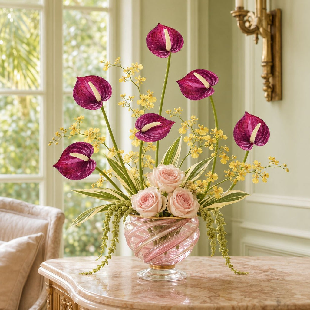 A floral arrangement in a pink glass vase on a marble table, featuring magenta anthuriums, pink roses, and small yellow flowers in a bright indoor room.
