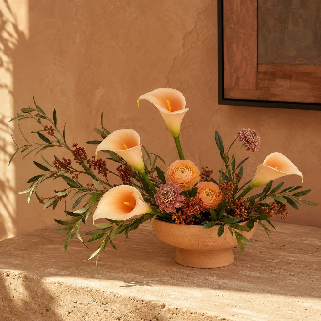 A terracotta flower arrangement featuring cream calla lilies, peach ranunculus, burgundy berries, and green foliage in a matching round pedestal vase, set on a stone surface against a warm stucco wall.