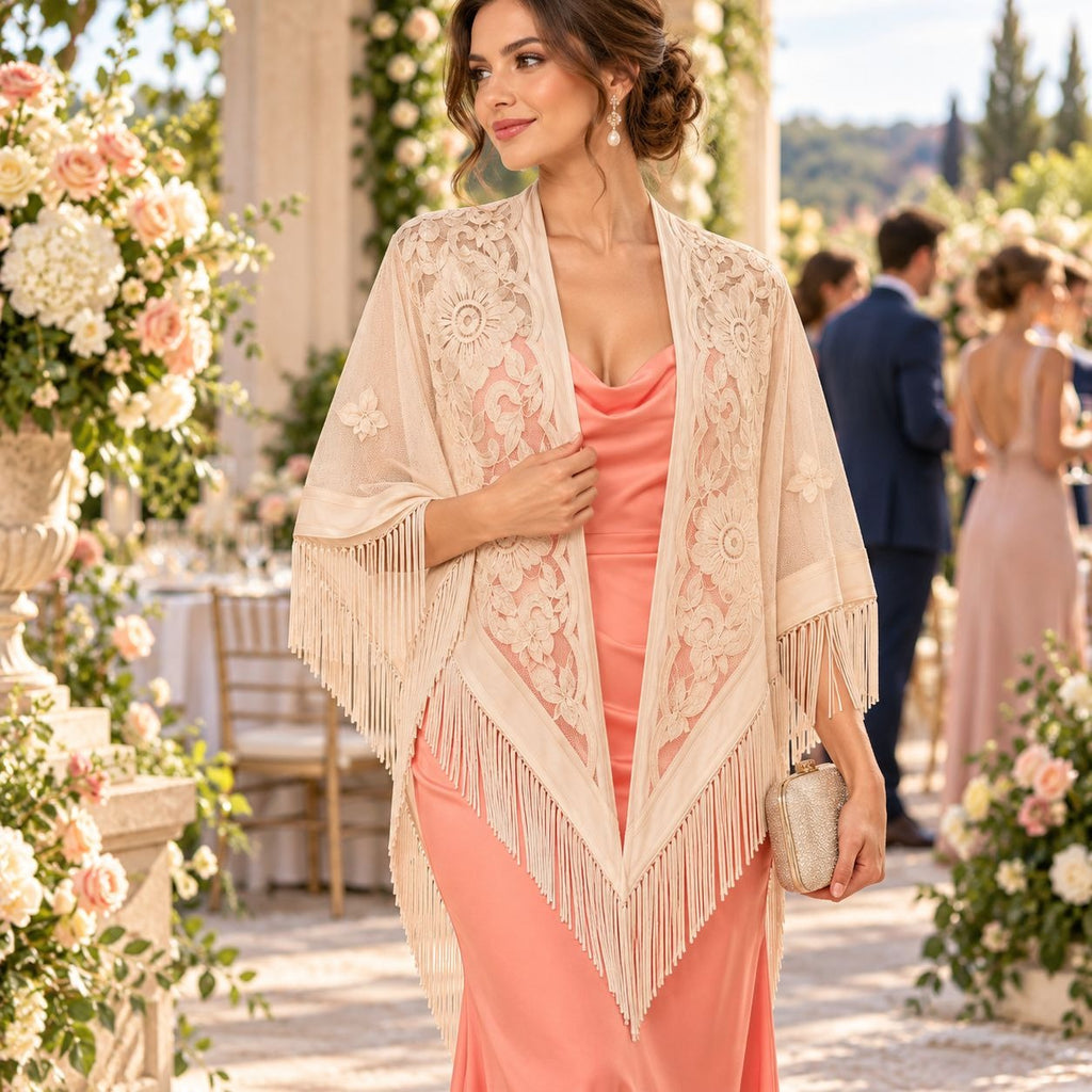 A woman outdoors wearing a peach pink satin slip dress and a cream lace shawl with fringe, holding a small metallic clutch. The background shows a sunlit garden event space with floral arrangements and blurred guests.