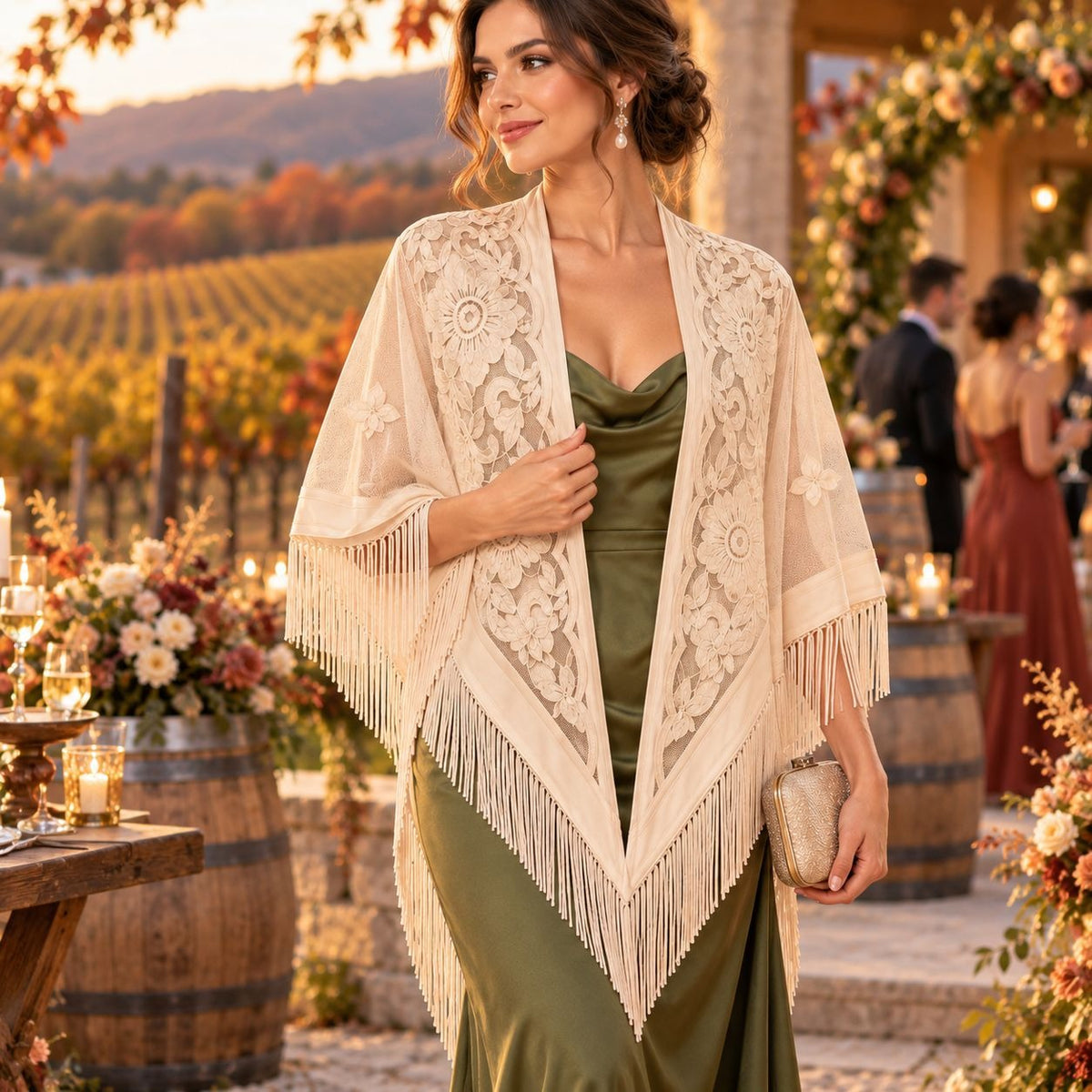 A woman stands outdoors in a vineyard setting wearing a cream lace shawl with fringe over an olive green dress, holding a metallic clutch.