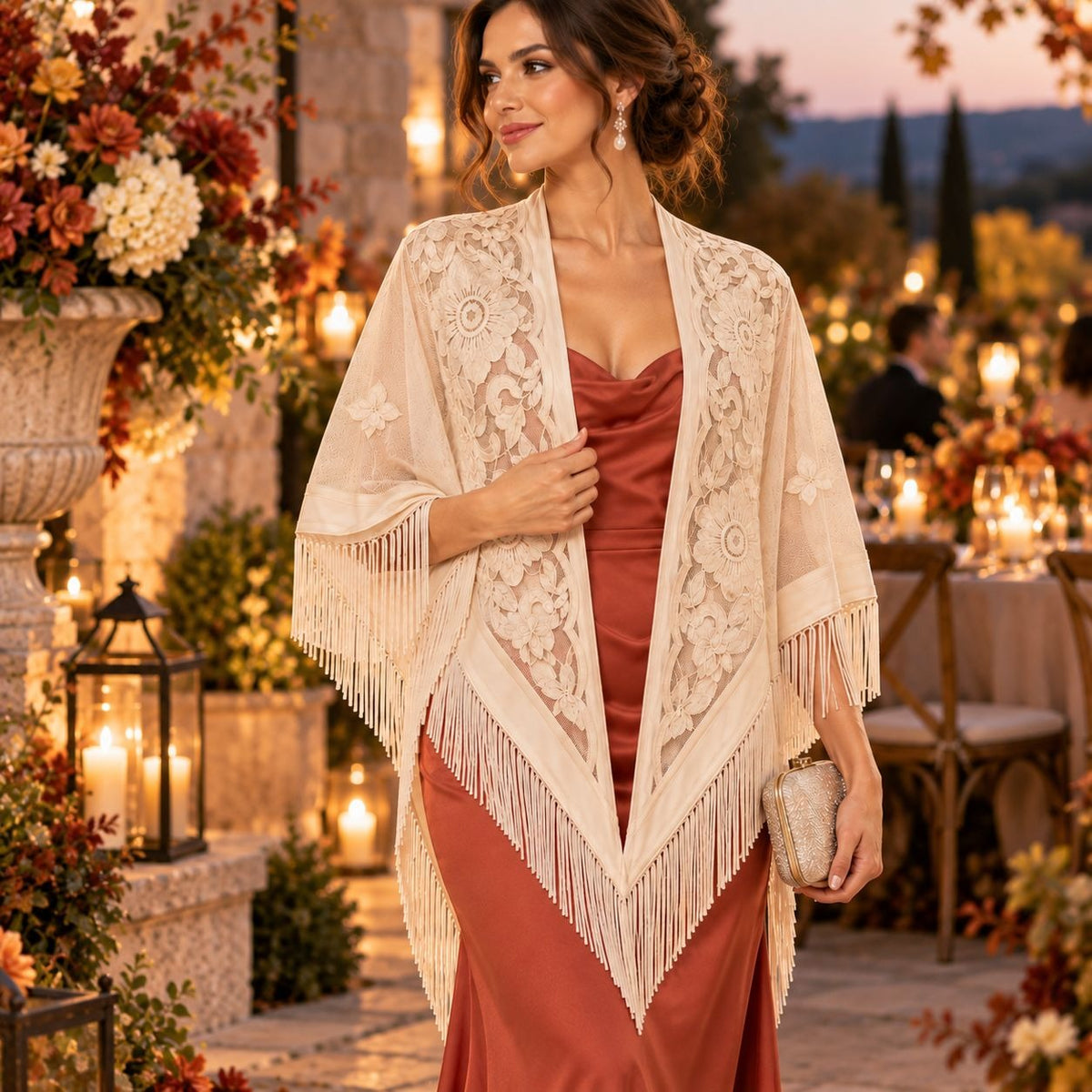 A woman stands outdoors at dusk wearing a cream lace shawl with fringe over a rust-colored satin dress. The setting includes warm lanterns, floral arrangements, and evening tables in the background.