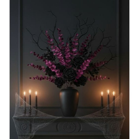 Gothic floral arrangement in a black vase on a mantel, featuring black roses, magenta blossoms, and bare branches, with lit candles and cobweb decor against a dark wall.