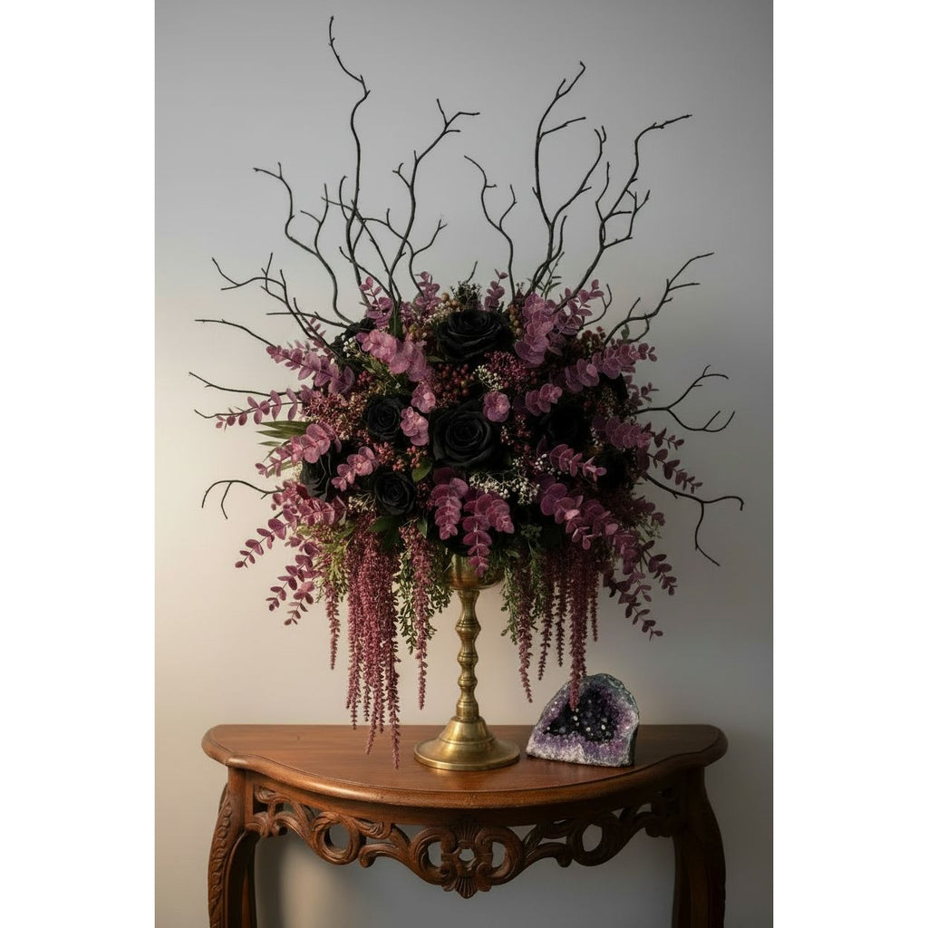 A large artificial floral arrangement in a gold pedestal vase, placed on an ornate wooden table against a plain light gray wall. The bouquet features black roses, pink and purple flowers, green foliage, and tall bare black branches extending upward.