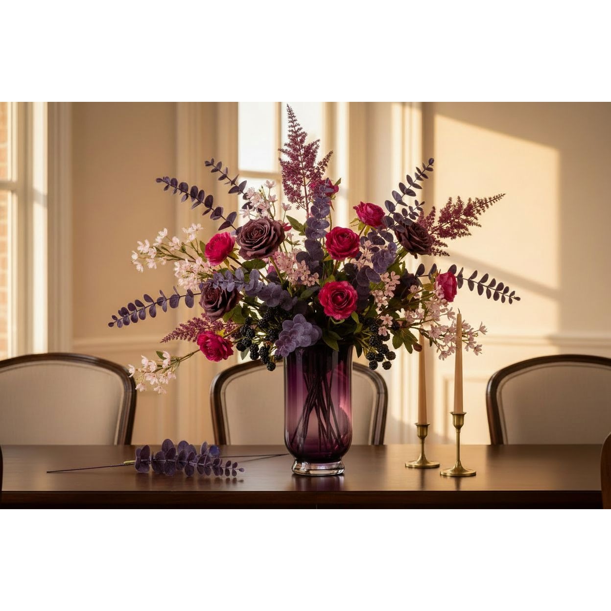 A purple glass vase filled with a floral arrangement of pink and purple roses, lavender, and green foliage, set on a dark wooden table in a warmly lit room with chairs and candles in the background.