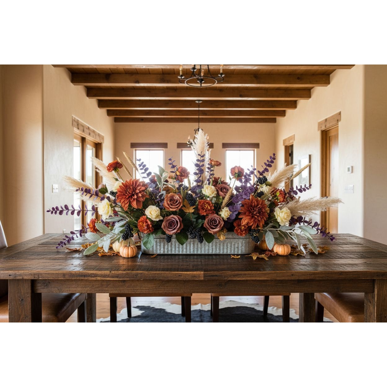 A large autumn floral centerpiece arranged in a galvanized metal rectangular planter on a dark wood dining table, set in a warm, rustic dining room with wooden ceiling beams.