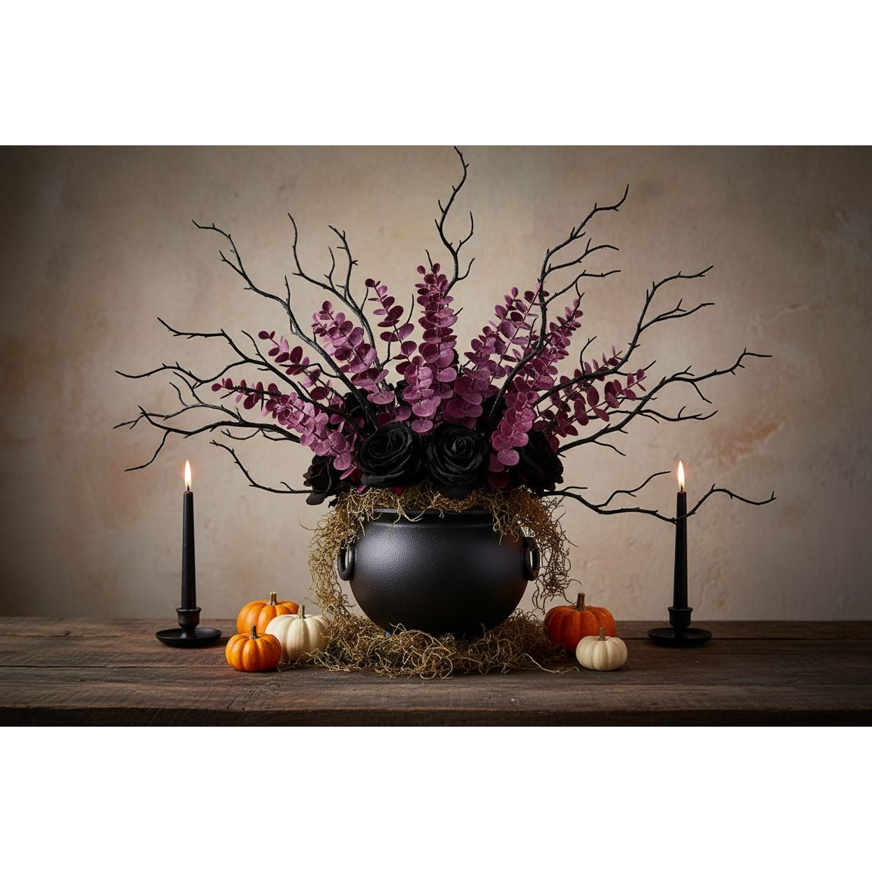 A gothic-style floral arrangement in a black cauldron vase with purple flowers, black roses, and bare branches, displayed on a rustic wooden table with lit black candles and small pumpkins against a textured beige backdrop.
